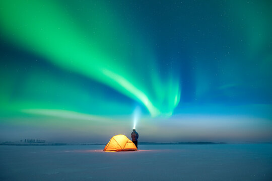 Tourist with flashlight near yellow tent against the backdrop of incredible starry sky with Aurora borealis. Gorgeous night landscape with polar lights. Northern lights in winter field