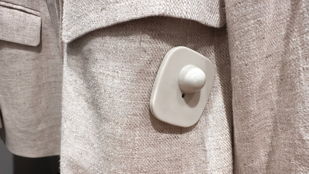 A close-up of a security tag attached to a beige fabric garment in a retail store.

