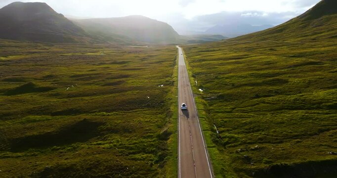 Drone follows white tourist car driving on famous NC500 with epic scenery of Scottish fields and hills on sunny day