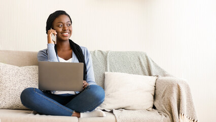 Black millennial woman using laptop and talking on cellphone while sitting on sofa at home, panorama with free space