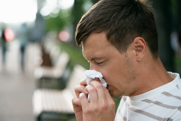 Close-up of a man blowing his nose into a tissue while sitting in a park, highlighting hygiene and...