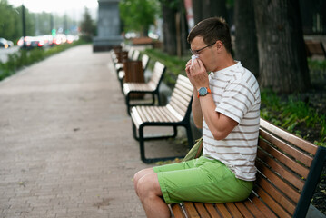 Obraz premium Seasonal allergies or a cold with a runny nose. Man wearing a striped shirt and green shorts sits on a bench in a park, sneezing into a tissue on a calm day.