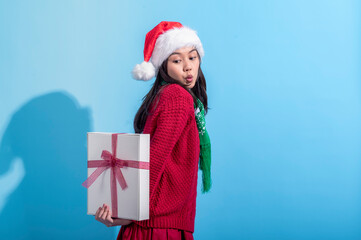 An Asian woman wearing a Santa hat, red sweater, and green scarf smiles playfully while holding a white gift box tied with a pink ribbon behind her back. She stands against a light blue background