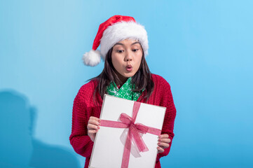 An Asian woman wearing a Santa hat, red sweater, and green scarf smiles warmly while holding a white gift box tied with a pink ribbon. She stands against a light blue background
