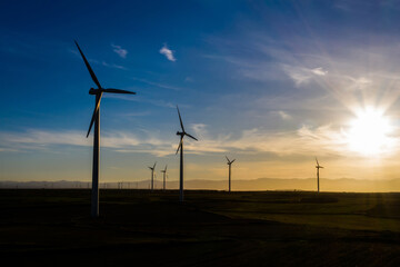 Obraz premium Silhouette Modern windmills with propellers standing in rural farmland agri meadow over orange sunset sky. Wind blowing generating renewable energy for clean resources. Electric power production
