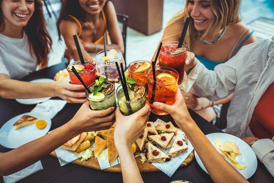 Happy female people toasting cocktail glasses sitting at bar table - Cheerful friends enjoying happy hour at pub restaurant balcony - Food and beverage life style concept