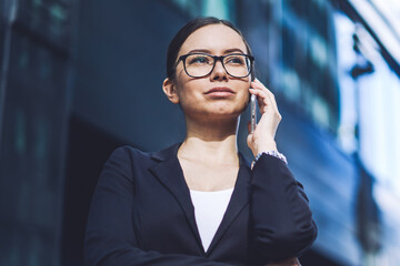 Serious business woman dressed in formal wear making concentrated smartphone conversation during break on urban setting, attractive confident female finance professional calling to colleague