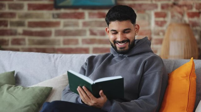 Positive hispanic male sitting on pillowed sofa and reading fascinating book during daytime at home. Young male enjoying useful hobby and getting interested by excited plot of fiction literature.