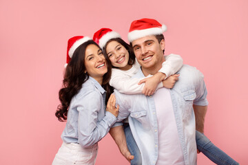 Adorable and happy European family posing in Santa hats, embracing and smiling at camera over pink studio background