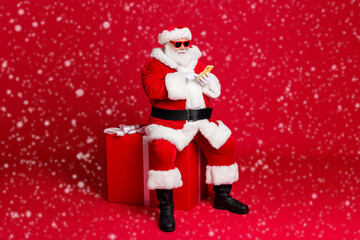 Full length photo of retired old man white beard hold telephone sit large box what chatting elf friend wear x-mas santa costume sunglass headwear leather boot isolated red color background