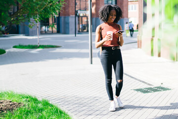 Millennial female student in casual wear walking on urban setting with cellular phone for online communication, positive dark skinned hipster girl writing sms message during coffee time outdoors