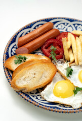 Breakfast plate with fried eggs, sausage, tomatoes, and fries served on a decorative blue plate