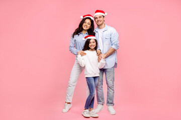 European family in Santa hats, hugging and smiling at camera, posing against pink background, full length shot. Celebrating winter holidays together