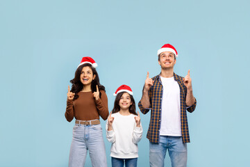 Excited European parents and girl wearing Santa hats and pointing up at copy space above their heads, family demonstrating place for Xmas advertisement, blue background