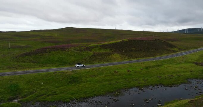 Cinematic aerial shot of white electro car driving with beautiful scenery of famous Highlands in Scotland
