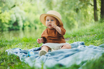 A little boy is sitting in the grass outdoors in the park with a hat and smiling. The child is sitting in the park on the grass. A boy in a straw hat.