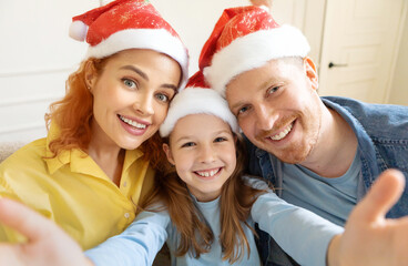 Happy European family of three in Santa hats taking selfie on phone at home, enjoy holiday and festive mood, mother, father and girl smiling at camera