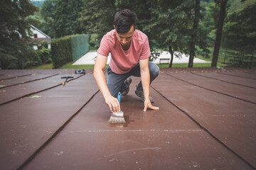 Focused man kneels on a roof while meticulously applying a fresh coat of paint with a wide brush....