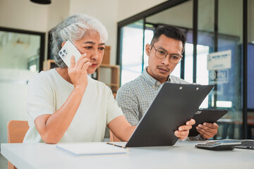 Fototapeta premium Senior female specialist talking on the phone, writing notes while new colleague is working on laptop, teamwork or multitasking, manager in meeting, financial accounting concept