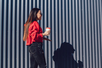 Side view of stylish hipster girl dressed in fashionable apparel waking around urban setting with caffeine beverage and modern smartphone in hand, chinese female teengaer holding coffee to go