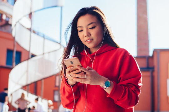Attractive chinese hipster girl writing mail via smartphone application while listening intersting audio book in electronics headphones, millennial woman installing app for creating music playlist