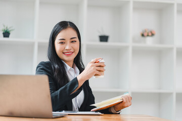 professional woman smiles confidently while holding coffee cup and notebook in modern office setting. bright and organized workspace enhances her positive demeanor.