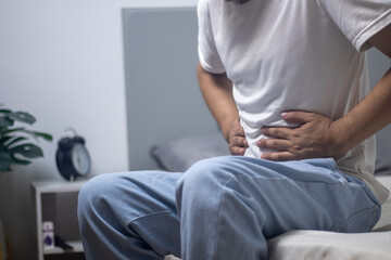 The man is sitting on the bed, clutching his stomach in pain.