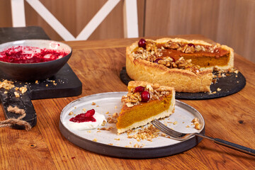 A slice of pumpkin pie with cream and cranberries rests on a ceramic plate on a wooden table