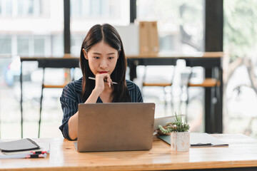 Focused woman working on laptop in modern office setting, deep in thought while holding pen....
