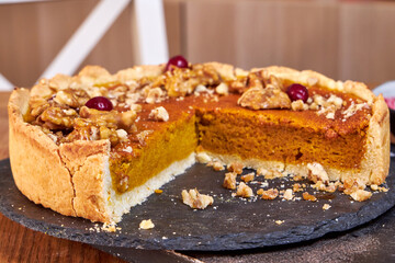 pumpkin pie decorated with nuts and cranberries stands on a black stand on a wooden table side view