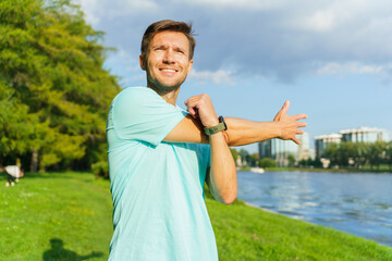 A Man Stretches His Arm by the Riverbank on a Sunny Day, Enjoying Outdoor Exercise in a Vibrant Green Park Setting