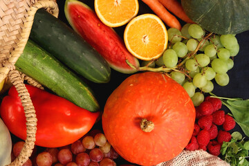 Round straw bag and various seasonal fruits and vegetables on dark background. Summer and fall produce. Top view.