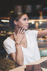 Attractive hipster girl dressed in casual look clicking selfie photos via cellular phone while sitting at table with modern laptop computer, charming woman taking pictures via application on cellphone