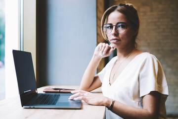 Portrait of attractive female programmer working remotely and looking at camera in break of creating new website, beautiful skilled woman in optical eyewear sitting at table with new modern netbook