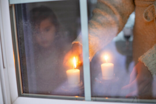 A cozy atmosphere as a person lights candles by a frosty window, with another person's reflection visible.