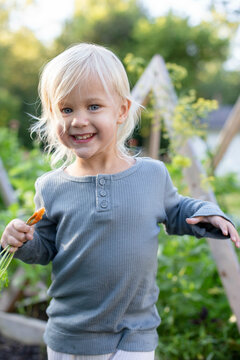 Smiling blonde toddler holding a carrot in a sunny garden setting.