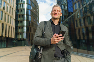 A Professional Man Smiles While Checking His Smartphone in a Modern Urban Area on a Sunny Day