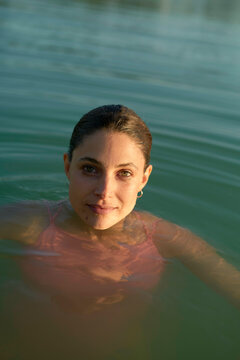 Woman bathing in a body of water with her hair pulled back and sunlight highlighting her face.