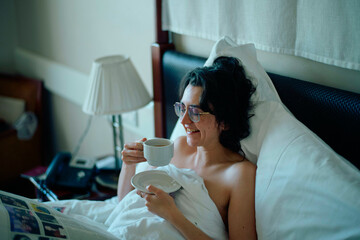 Adult woman enjoys a cup of coffee while reading a magazine in bed.