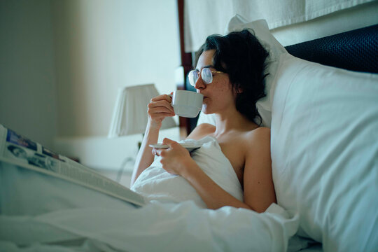 A woman relaxes in bed with a cup of coffee and a newspaper in a cozy bedroom setting.