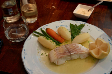 A meal of poached fish with lemon, boiled potatoes, carrots, and spinach served on a white plate with a glass of water and white wine on a wooden table.