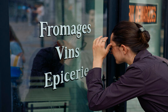 A woman intently looks through a shop window with text advertising cheese, wine, and groceries.