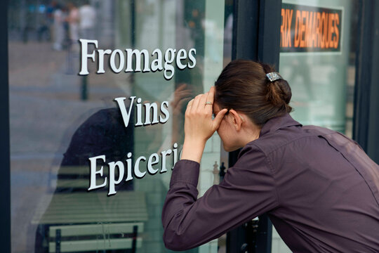A stressed adult woman holds her head in front of a shop window with 'Fromages Vins Epicerie' lettering.