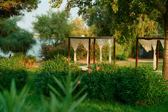 Sunrise light bathes a serene garden with a gazebo and lake in the background, embraced by rich green foliage and flowering plants.