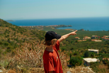 A person in a red shirt and black cap points towards the sea from a scenic overlook with lush greenery.