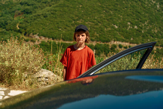 A child in a red shirt and black cap stands by a car door with a lollipop in their mouth, overlooking a lush green hillside.