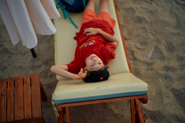 Child in a red dress lying on a beach lounge chair, smiling and waving at the camera.