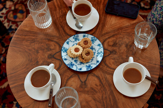 Overhead view of a wooden table with three cups of coffee, a plate of cookies, glasses of water, and a smartphone, with partial views of two people.