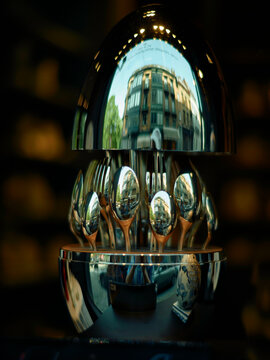 A reflective silver sphere distorts the image of a cityscape, with a set of spoons and forks arranged in a circular pattern around the base.