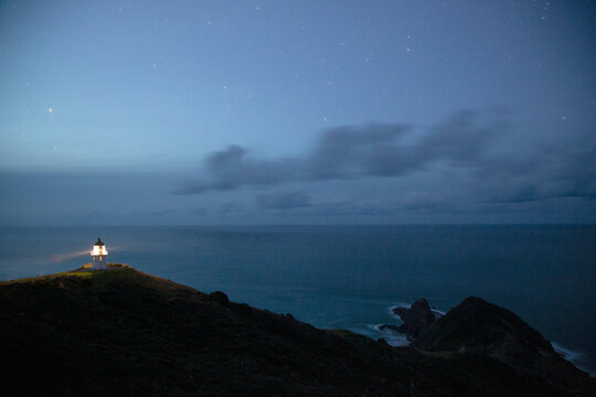 A lighthouse illuminates the night sky over a rugged coastline under a starry sky, Cape Reinga, Northland, New Zealand